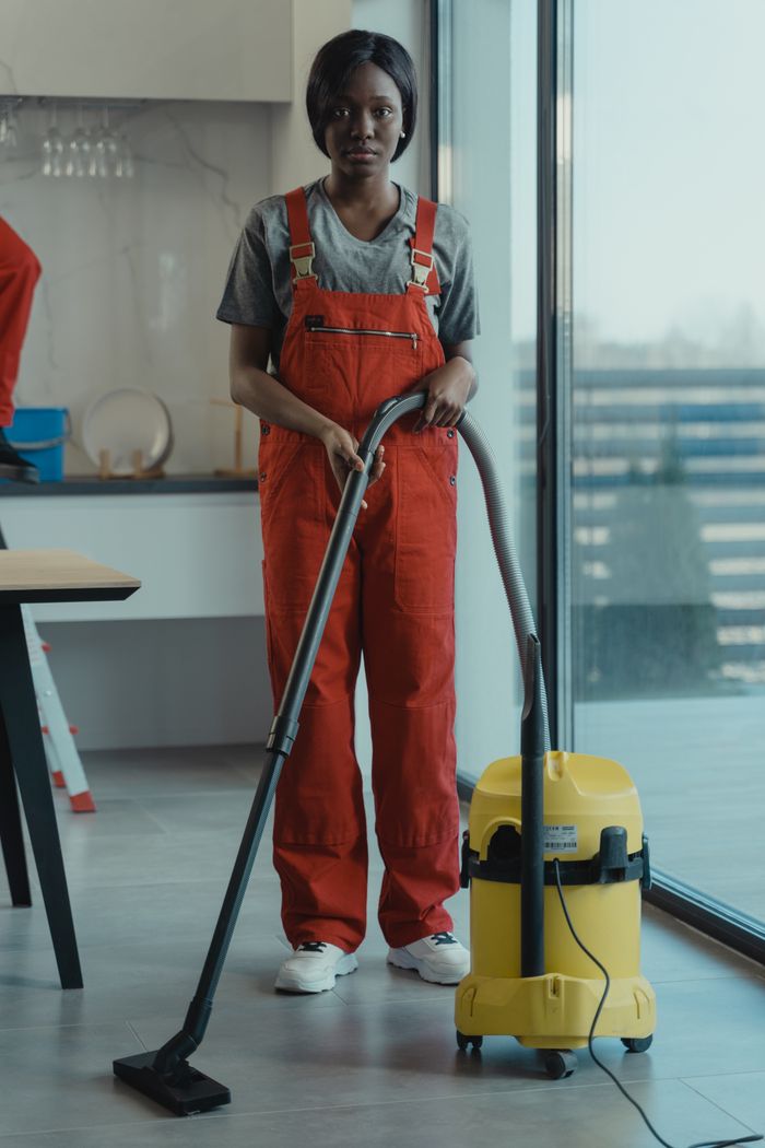 Une femme adulte vêtue d'une combinaison rouge utilise un aspirateur dans un espace intérieur contemporain, concentrée sur ses tâches de nettoyage.